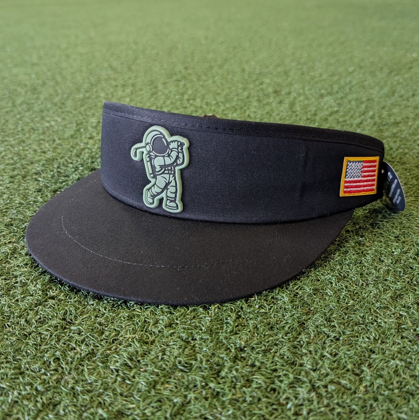 Black golf visor with glow in the dark astronaut design and American flag patch on green grass
